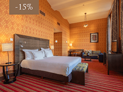 Een ruime hotelkamer met een klassieke uitstraling, inclusief een comfortabel tweepersoonsbed, kleurrijke tapijten en stijlvolle meubels. Een 15% korting wordt aangegeven in de linkerbovenhoek.