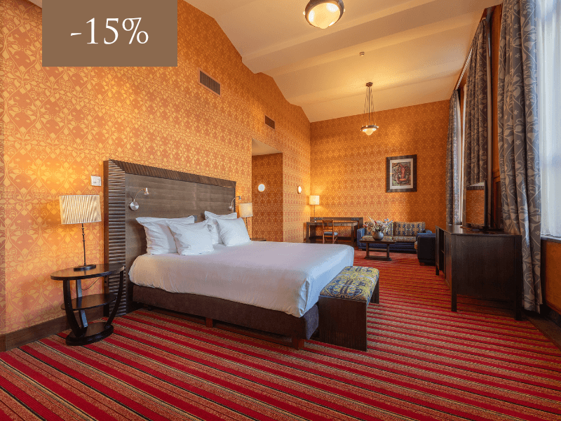 Een ruime hotelkamer met een klassieke uitstraling, inclusief een comfortabel tweepersoonsbed, kleurrijke tapijten en stijlvolle meubels. Een 15% korting wordt aangegeven in de linkerbovenhoek.