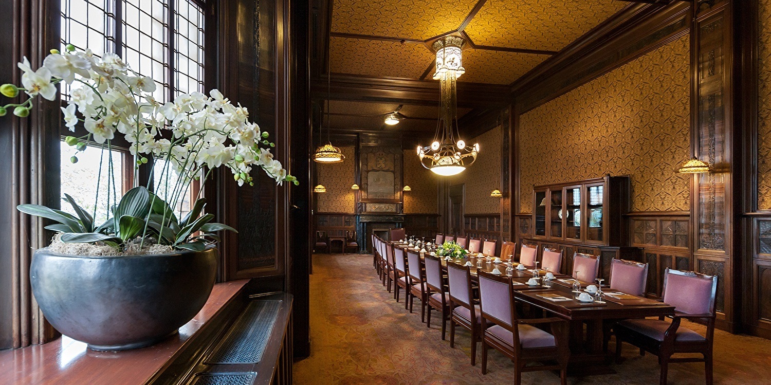 The Beraadzaal at Grand Hotel Amrâth Amsterdam, a luxurious meeting room featuring a long set table surrounded by elegant chairs, warm wooden paneling, and atmospheric chandeliers. In the foreground, a large pot of white orchids sits by a window.