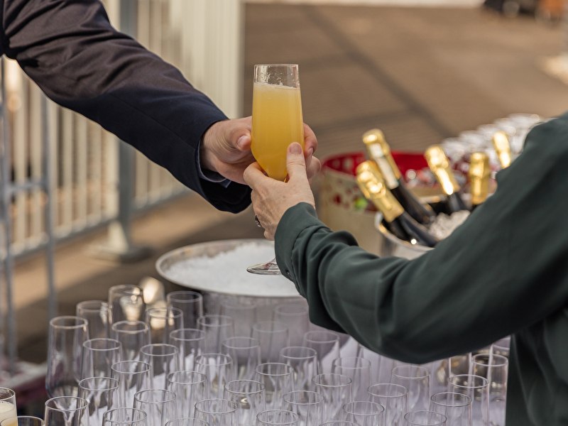 Gast ontvangt een glas mimosa van een medewerker op het dakterras van Grand Hotel Amrâth Amsterdam, met champagneflessen op ijs.