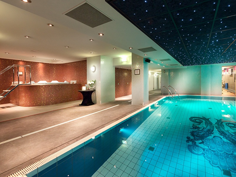 Een luxe wellnessruimte met een verlicht zwembad en een plafond met sterrenlichteffect. Links bevindt zich een jacuzzi-ruimte met mozaïektegels, en op de achtergrond zijn verschillende faciliteiten zichtbaar.