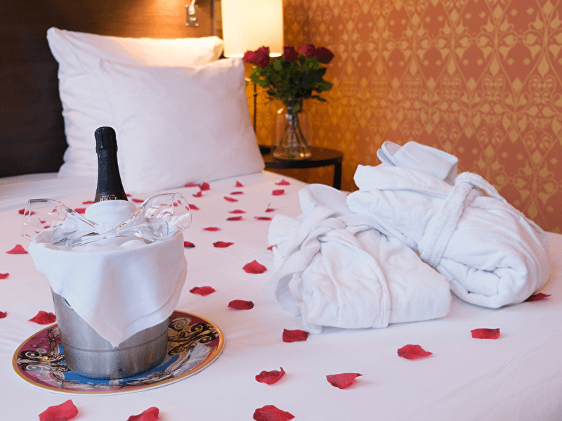 Een romantisch opgemaakt bed met rozenblaadjes, een gekoelde fles champagne in een emmer, twee glazen en twee witte badjassen. Op de achtergrond staat een vaas met rode rozen.