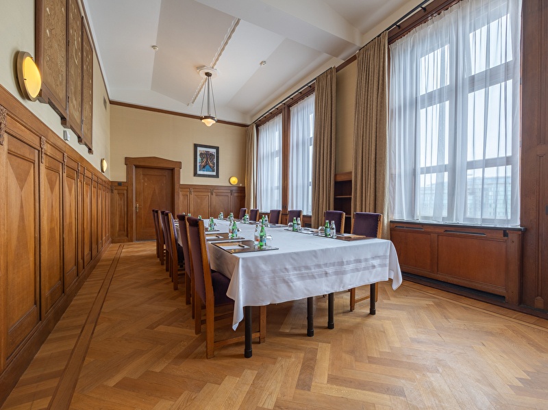 Stilvoller Tagungsraum im Grand Hotel Amrâth Amsterdam mit Holzvertäfelung, einem langen Tisch mit weißer Tischdecke, lila Stühlen und großen Fenstern für natürliches Licht.