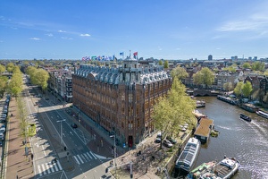 Schuin vogelperspectief van Grand Hotel Amrâth Amsterdam, gelegen langs een gracht met boten en omringd door historische gebouwen en een levendige stadsomgeving.